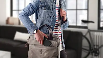 Eine Person in Jeansjacke verstaut ein schwarzes Gerät in einer grauen Umhängetasche. Im Hintergrund sind Fenster und ein Fahrrad.