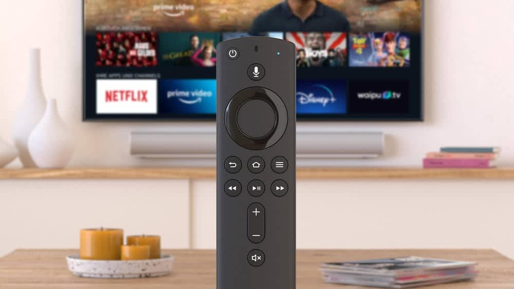 Eine Amazon-Fire-TV-Stick-Fernbedienung steht auf einem Tisch, im Hintergrund läuft ein Fernseher mit Amazon Fire TV.