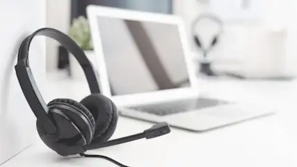 Schwarzes Headset mit Mikrofon vor einem weißen Laptop auf einem weißen Tisch. Ein weiteres Headset ist im Hintergrund zu sehen.