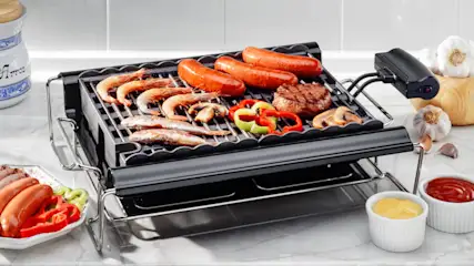 Ein Tischgrill mit Würstchen, Garnelen, Pilzen, Paprika und einem Steak. Saucen, Knoblauch und Geschirr sind auch sichtbar.