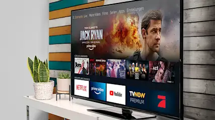 Ein Flachbildfernseher zeigt Streaming-Apps wie Netflix, YouTube und Jack Ryan vor einer Wand mit Holzpaneelen. Zimmerpflanzen stehen auf einem Tisch.