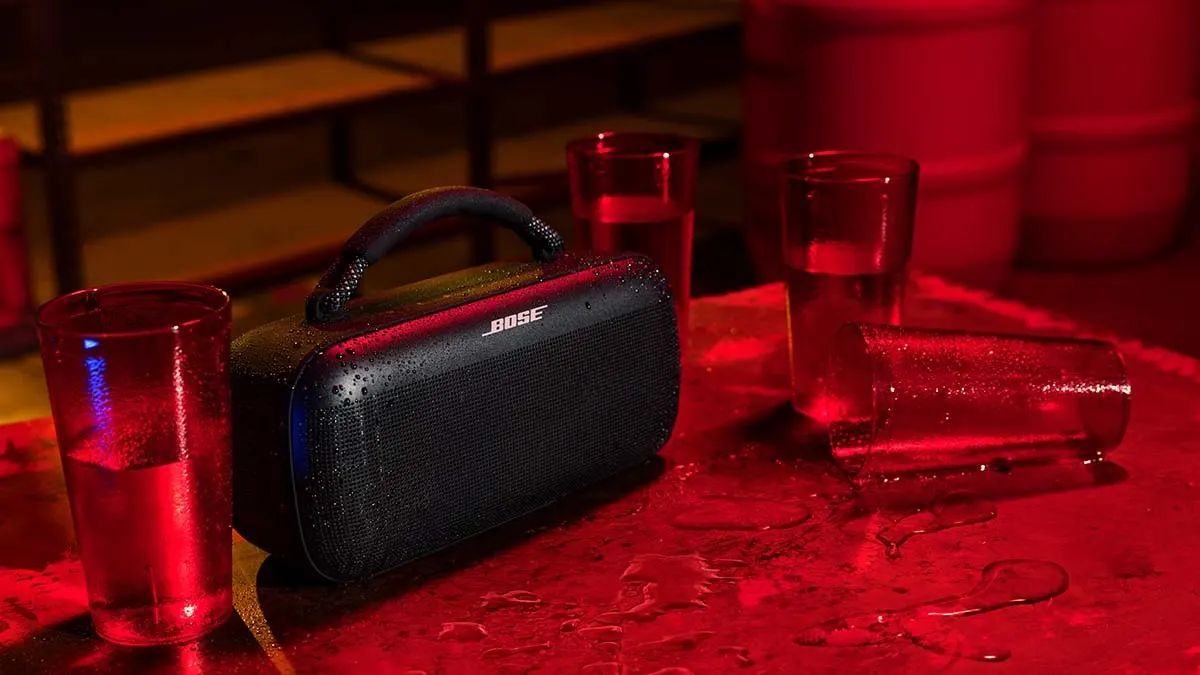 Ein schwarzer Bose-Lautsprecher steht auf einem roten Tisch, umgeben von Gläsern mit Flüssigkeit. Eines der Gläser liegt umgekippt.