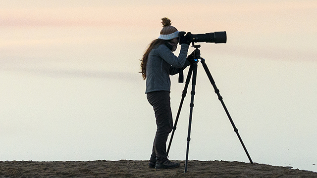 Eine Person mit Mütze bedient eine Kamera auf einem Stativ im Freien vor einem hellen Hintergrund.