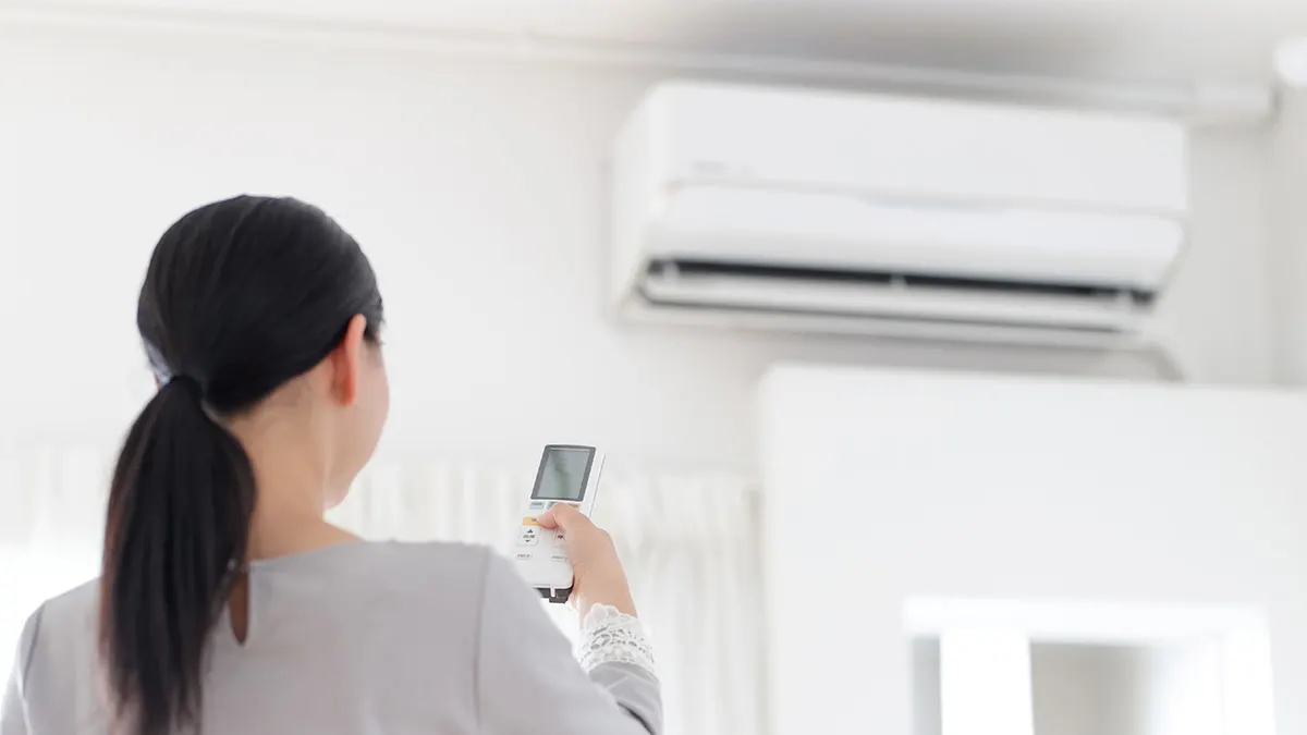 Eine Frau ist von hinten zu sehen. Sie bedient mit einer Fernbedienung eine Klimaanlage an der Wand in einem hellen Raum.