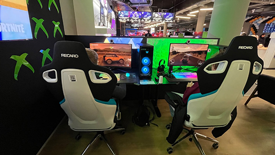 Zwei Recaro-Gaming-Stühle stehen vor Computern mit Rennspielen, flankiert von grünen Xbox-Logos und Neonlichtern.