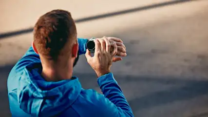 Ein Mann in blauer Jacke schaut auf eine schwarze Smartwatch am Handgelenk, Hintergrund ist verschwommen.