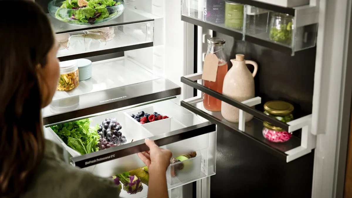 Ein Kühlschrank ist mit Salaten, Beeren, Gurken und Getränken gefüllt. Eine Person öffnet eine Schublade.