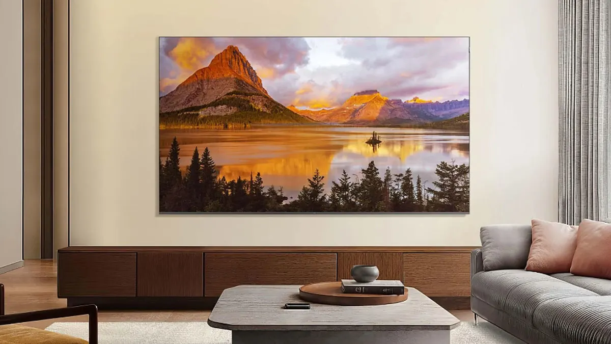 Der Fernseher TCL85P8K zeigt ein Landschaftsbild. Er hängt an einer Wohnzimmerwand, davor steht ein grauer Couchtisch.