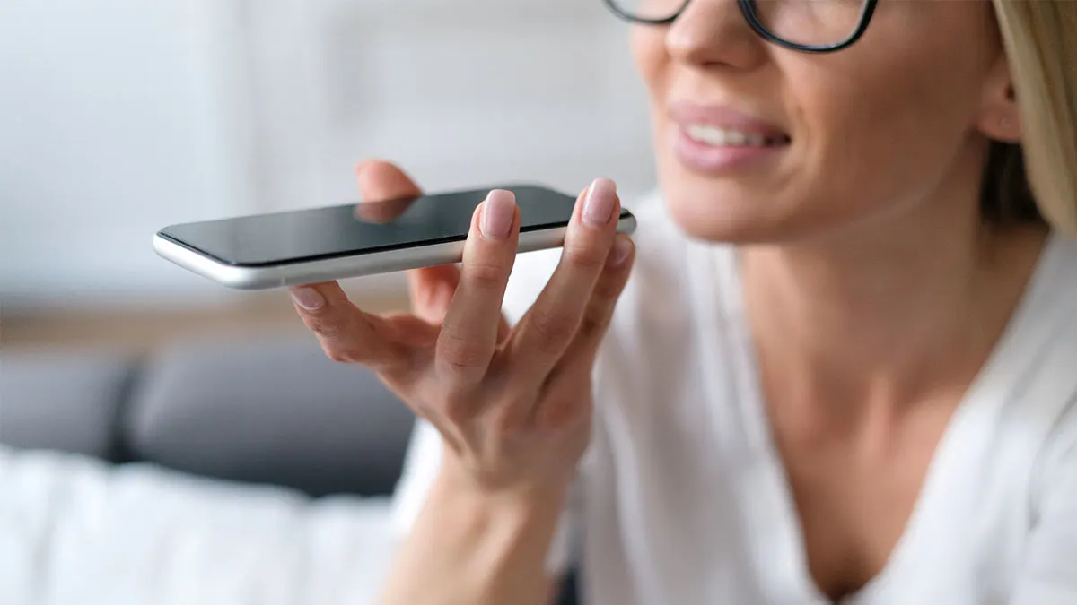 Blonde Frau mit Brille hält ein Smartphone vor den Mund und spricht hinein.