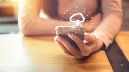 Eine Person hält ein Smartphone mit einer Cloud-Grafik und einem Download-Pfeil darüber, auf einem Holztisch.