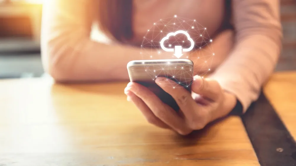 Eine Person hält ein Smartphone mit einer Cloud-Grafik und einem Pfeil nach unten über einem Holztisch.