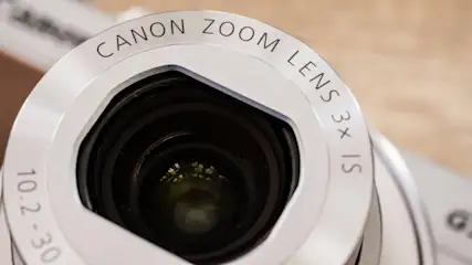 Nahaufnahme eines silbernen Canon Zoomobjektivs mit schwarzer Linse, Details wie "3x IS" und "10.2-30" sind sichtbar.