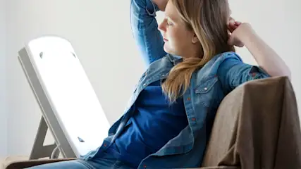 Frau in weißem Hemd mit geschlossenen Augen vor hellem Lichtpanel auf grauem Hintergrund.