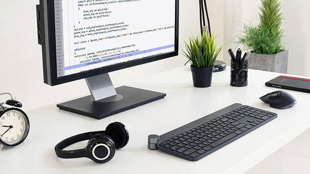 Ein Schreibtisch mit einem Monitor, einer Tastatur, einer Maus, Kopfhörern, einer Uhr, Stiften, Pflanzen und einem Notizbuch.