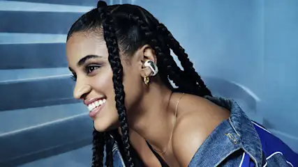 Eine lächelnde Person mit Zöpfen trägt einen silbernen Ohrhörer und einen goldenen Ohrring vor einem blauen Hintergrund.