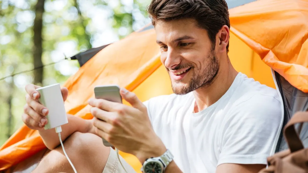 Ein Mann sitzt vor einem Zelt und lädt sein Smartphone mit einer Powerbank.