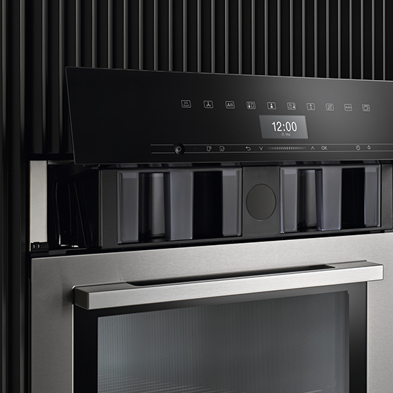 Nahaufnahme eines modernen, eingebauten Küchengeräts mit digitaler Anzeige "12:00" und Edelstahlgriffen.