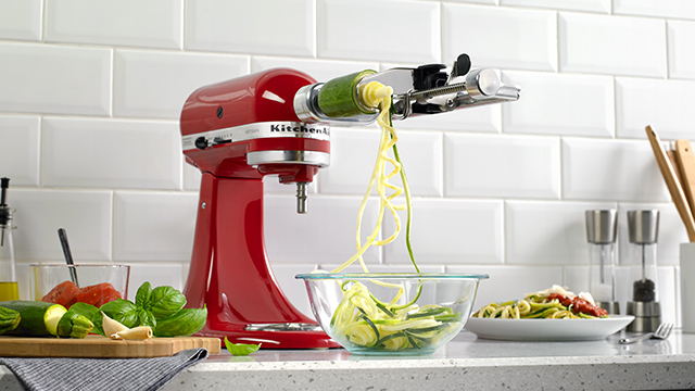 Eine rote Küchenmaschine spiralisiert eine Zucchini in eine Glasschüssel auf einer Küchentheke. Gemüse liegt daneben.