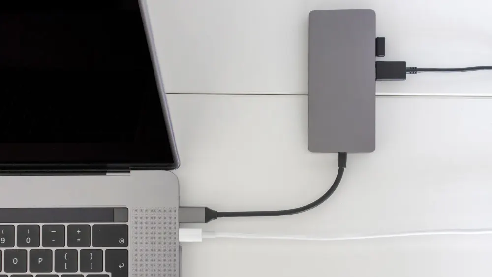 Ein grauer Laptop ist mit einem Hub verbunden, der an einer weißen Wand befestigt ist und über schwarze Kabel weitere Geräte verbindet.