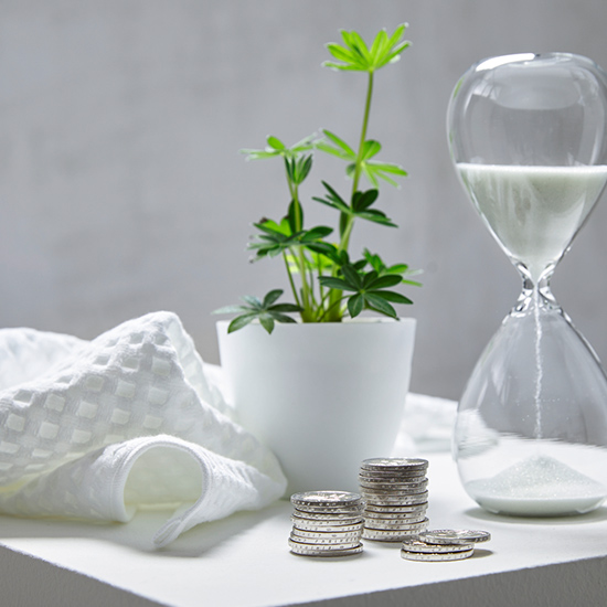 Eine weiße Decke, Geldstapel, eine Pflanze in einem weißen Topf und eine Sanduhr stehen auf einer weißen Oberfläche.
