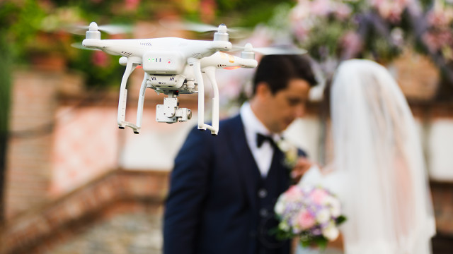 Eine weiße Drohne schwebt vor einem Mann im Anzug mit Fliege und einer Braut mit Schleier, beide halten Blumen.