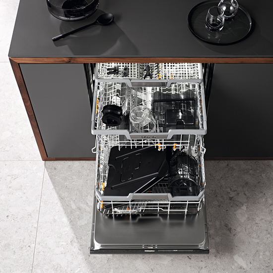 Ein offener Geschirrspüler mit zwei beladenen Körben unter einer dunklen Arbeitsplatte in einer Küche. Geschirr und Gläser sind sichtbar.