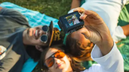 Drei Personen liegen auf einer Decke im Gras. Eine Hand hält eine schwarze Kamera, die ein Selfie der Gruppe zeigt.
