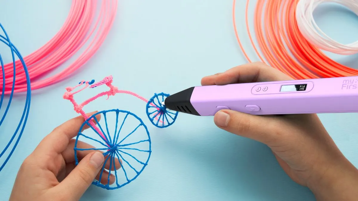 Eine Person benutzt einen lila 3D-Stift, um ein blaues und pinkes Fahrrad auf einem blauen Untergrund zu erstellen. Filamentrollen im Hintergrund.