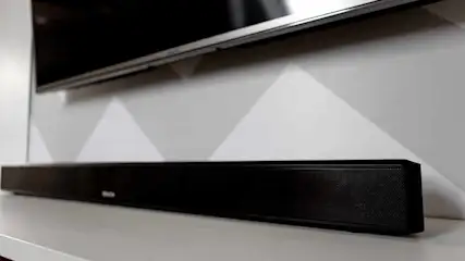 Ein schwarzer Fernseher ist über einer Soundbar auf einem weißen Tisch montiert, vor einer Wand mit dreieckigem Muster.