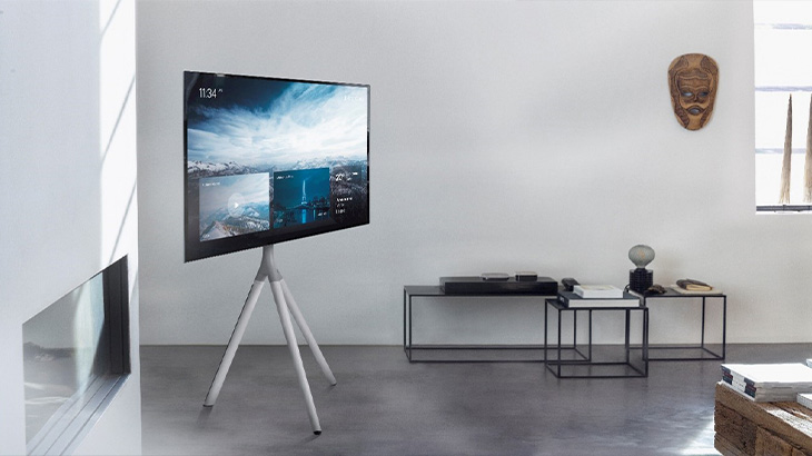 Ein Zimmer mit einem schwarzen Fernseher auf einem weißen Stativ. An der Wand hängt eine hölzerne Maske.