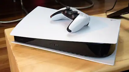 Auf einem Holztisch liegt eine weiße und schwarze PlayStation 5 mit einem weißen Controller darauf.