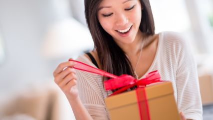Eine brünette Frau hält ein Geschenk in der Hand, auf das sie schaut. Mit der rechten Hand zieht sie das rote Geschenkband auf und lächelt vorfreudig.