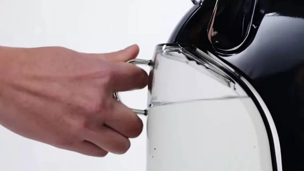 Eine Hand hält einen transparenten Krug mit einem silbernen Griff, während Wasser aus einem schwarzen Behälter eingegossen wird.