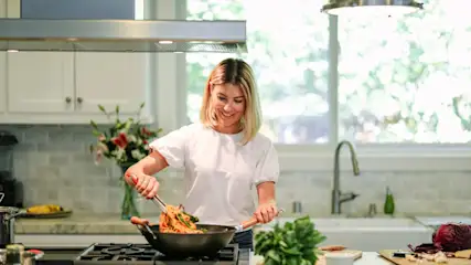 Eine Frau kocht in einer Küche mit weißen Schränken und einer Dunstabzugshaube über dem Herd, auf dem eine Wokpfanne steht.