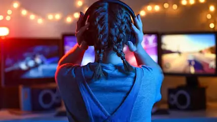 Person von hinten, trägt Kopfhörer, vor drei Monitoren mit Spielgrafiken. Lichterkette im Hintergrund.