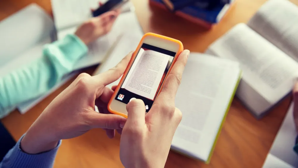 Eine Person fotografiert eine Buchseite mit einem orangefarbenen Smartphone. Auf dem Tisch liegen weitere Bücher und Papier.