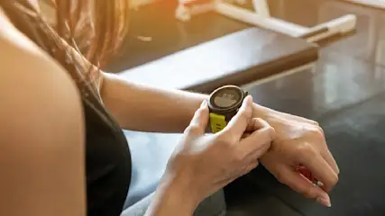 Eine Person betrachtet eine schwarze Smartwatch mit grünem Armband am Handgelenk vor einem Fitnessstudio-Hintergrund.