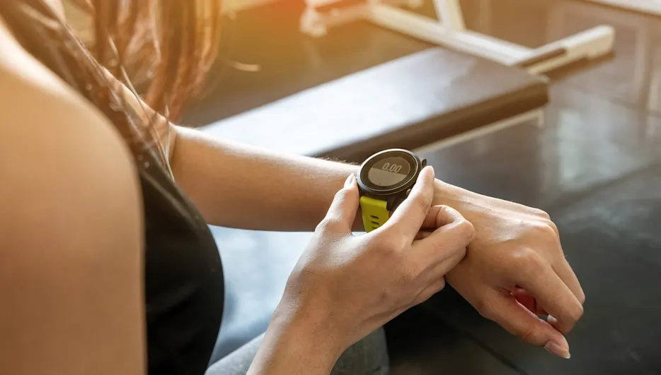 Eine Person betrachtet eine schwarze Smartwatch mit grünem Armband am Handgelenk vor einem Fitnessstudio-Hintergrund.