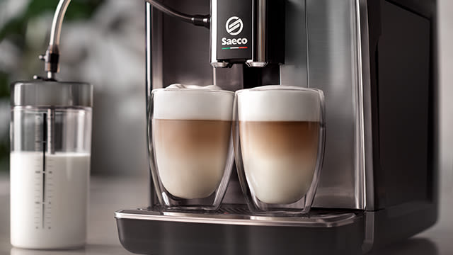 Ein Saeco Kaffeevollautomat unter welchem zwei gefüllte Tassen mit Latte Macchiato und daneben ein Behälter mit Milch steht.