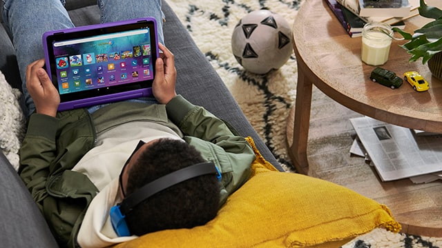 Eine Person mit Kopfhörern liegt auf einem Sofa und hält ein violettes Tablet. Ein Ball und ein Tisch mit Spielzeugautos sind im Hintergrund.