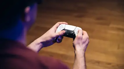Eine Person hält einen weißen und schwarzen Controller vor einem braunen Holzhintergrund. Die Person trägt ein dunkles Hemd.