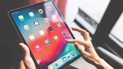 Eine Person bedient ein Tablet mit bunten App-Symbolen auf dem Bildschirm. Die Hand berührt das Display vor einem dunklen Hintergrund.