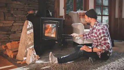Mann sitzt auf einem Teppich und stochert mit einem Schürhaken in einem schwarzen Ofen, daneben Steine und Holz.
