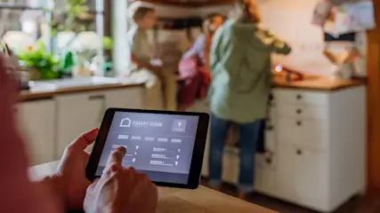 Ein Tablet zeigt eine Smart-Home-App, während eine Familie in einer Küche im Hintergrund steht.
