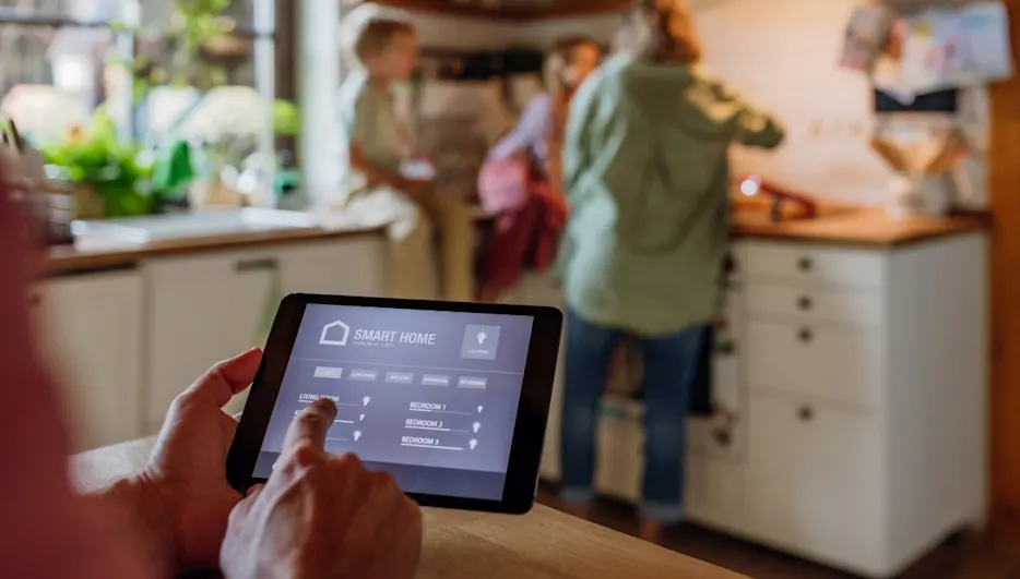 Ein Tablet zeigt eine Smart-Home-App, während eine Familie in einer Küche im Hintergrund steht.