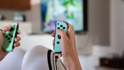 Hände halten grüne und blaue Controller vor einem verschwommenen Fernsehbildschirm. Eine Person sitzt auf einem weißen Sessel.