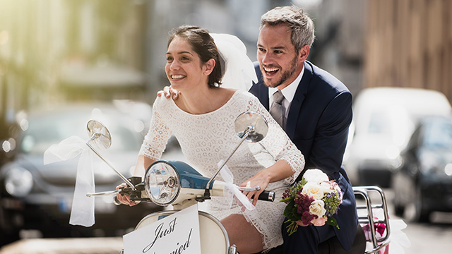 Ein Paar in Hochzeitskleidung fährt lachend auf einem hellblauen Motorroller mit Blumenschmuck durch eine Stadt.