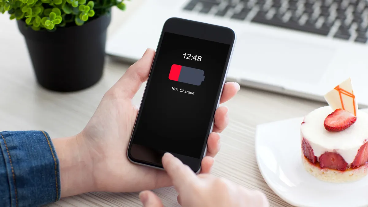 Auf dem Bildschirm eines Telefons wird eine Warnmeldung über einen niedrigen Batteriestand angezeigt. Dahinter ist eine Pflanze, ein Laptop und ein Erdbeertörtchen zu sehen.