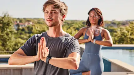 Ein Mann und eine Frau praktizieren Yoga im Freien, mit gefalteten Händen vor der Brust, vor Bäumen und unter blauem Himmel.