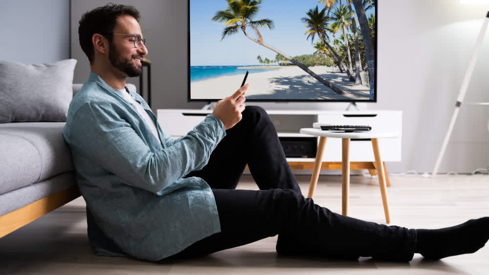 Ein Mann sitzt vor einem Fernseher auf dem Boden und blickt lachend auf sein Smartphone.